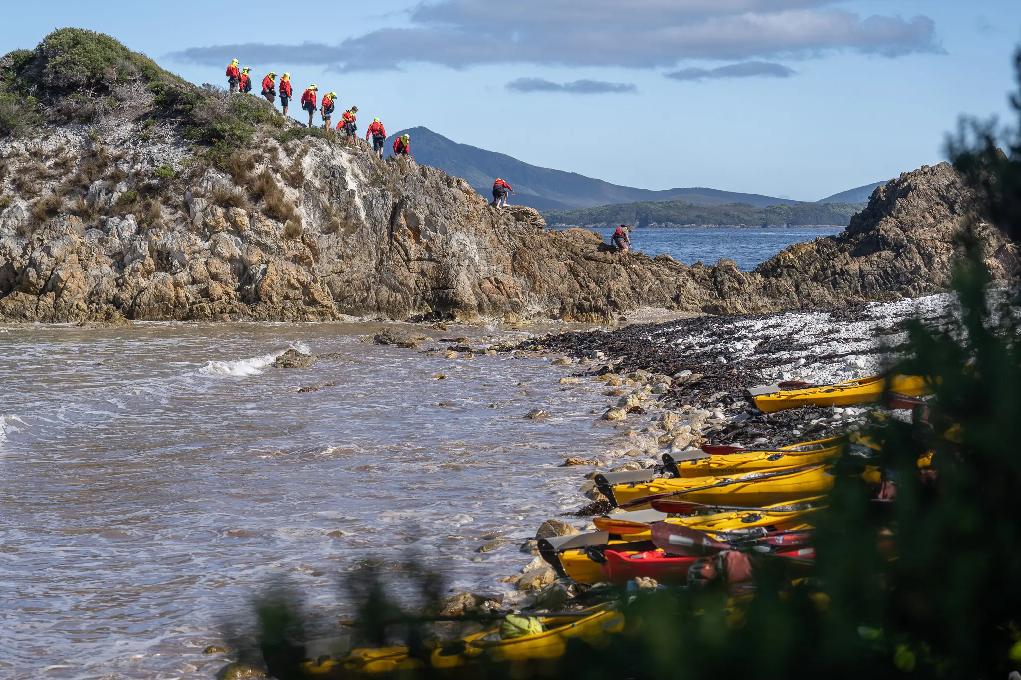 Hutchins Power of 9 students participating in the Port Davey Challenge climb on rocky coastal headlands while their kayaks rest on the beach below.